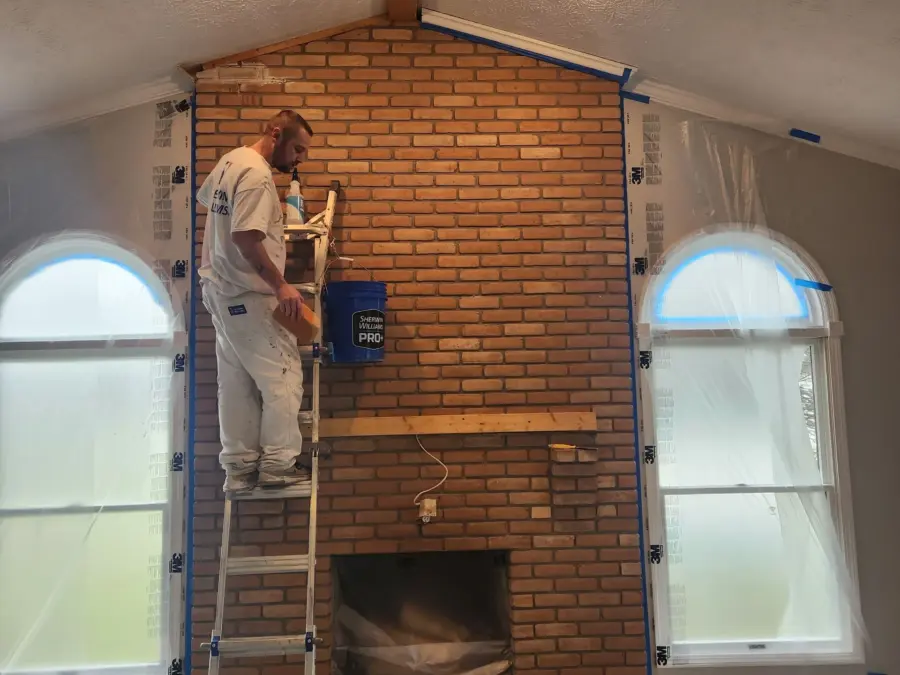 A man is standing on a ladder painting a brick fireplace in a living room.