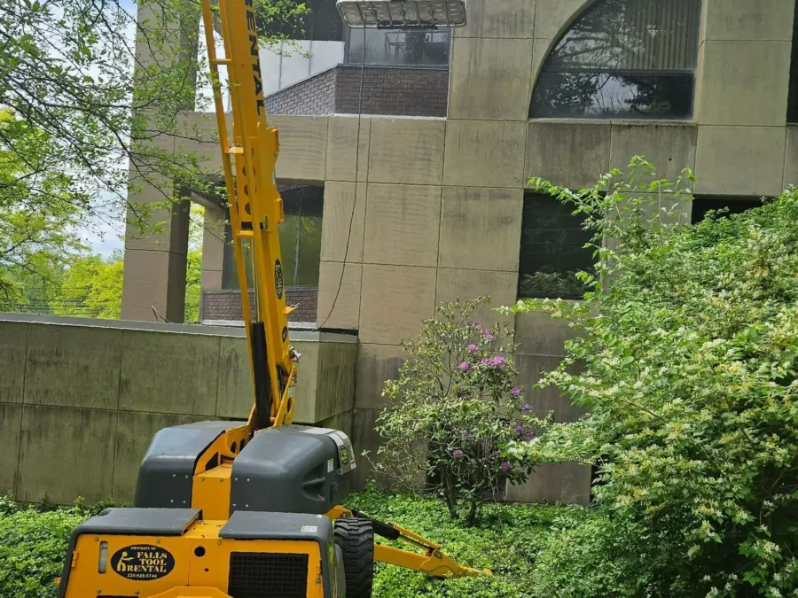A yellow tractor is cutting a tree in front of a building