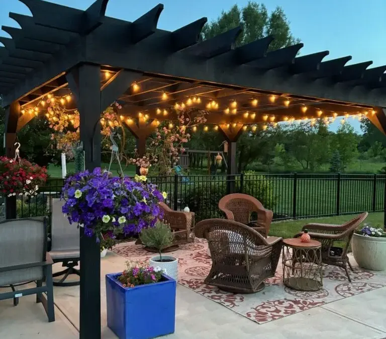 A patio with a pergola , chairs , tables and purple flowers.